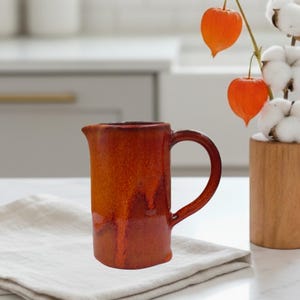 Puede incluir: Jarra de cerámica hecha a mano con un rico esmalte rojo-naranja. La jarra tiene una base cuadrada, un asa curva y un pico vertedor. Se encuentra sobre una servilleta de lino blanco, con flores naranjas decorativas y tallos de algodón en el fondo.