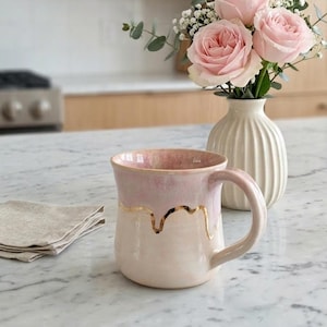 May include: A ceramic mug with a pink interior and a white exterior, decorated with gold accents. The mug is next to a small white vase holding pink roses. A folded beige linen napkin rests on the counter.