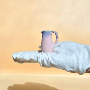 Può includere: Una brocca in ceramica in miniatura con un corpo liscio rosa chiaro che sfuma in uno smalto blu tenue attorno al bordo e al manico. Il piccolo recipiente presenta un manico curvo e un beccuccio semplice con una finitura lucida.