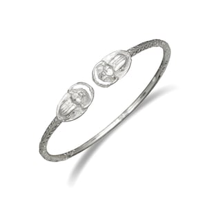 Könnte beinhalten: Ein silbernes Armreif-Armband mit zwei Skarabäus-Anhängern. Das Armband ist offen und hat eine strukturierte Oberfläche.