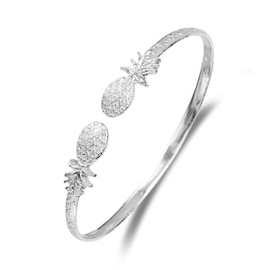 May include: A silver-toned open cuff bracelet featuring two pineapple-shaped charms at each end. The pineapple motifs have a textured, cross-hatched surface detail, and the band itself has a smooth, polished finish that reflects light.
