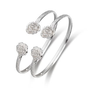 May include: Two silver-colored, open-ended bangles. Each bangle features a floral rose design at the ends. The bands have a textured pattern near the rose. The roses are detailed with multiple petals.