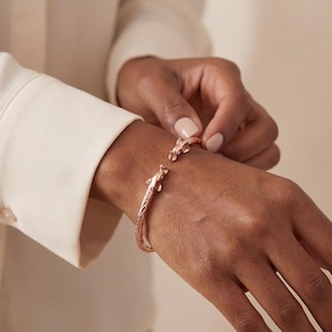 Könnte beinhalten: Roségoldenes Armreif-Armband mit zwei Alligatorköpfen. Das Armband hat ein strukturiertes Design und wird am Handgelenk geschlossen. Die Alligatorköpfe verleihen dem Schmuck eine einzigartige Note.
