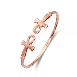 Könnte beinhalten: Roségoldfarbenes Armreif-Armband mit offenem Design und zwei Ankh-Symbolen. Das Armband hat ein strukturiertes Band, wahrscheinlich aus Metall. Die Ankh-Symbole sind ein Hauptmerkmal und verleihen eine altägyptische Ästhetik.