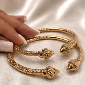 May include: Two gold-toned open cuff bracelets featuring a textured, engraved pattern along the band and pointed, ribbed finials at each end. The metal surface has a polished finish, and the design is circular with a gap for adjustment.