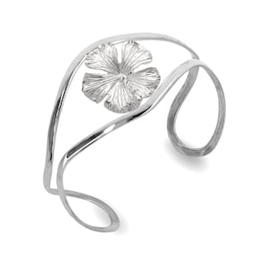 Könnte beinhalten: Ein silberfarbenes Armreif-Armband mit floralem Design. Das offene Armband hat eine stilisierte Blume in der Mitte, mit geschwungenen, fließenden Linien. Das Metall hat eine polierte Oberfläche, die das Licht reflektiert. Ein modernes, elegantes Schmuckstück.