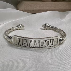 Könnte beinhalten: Ein silberfarbenes Armband mit einer zentralen rechteckigen Platte, auf der der Name MAMADOU in erhabenen Blockbuchstaben steht. Das Band weist ein strukturiertes, graviertes Muster auf und endet in abgerundeten, dekorativen Endstücken.