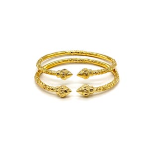 May include: Two gold-toned open cuff bracelets featuring a textured, patterned surface along the band and pointed, cone-shaped finials at each end. The bracelets are stacked one above the other, showcasing a polished, metallic finish throughout.