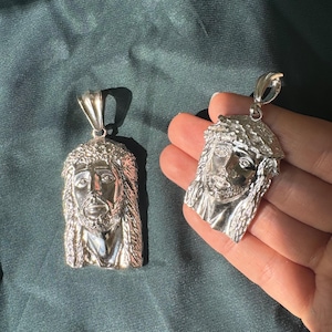 May include: Two silver pendants featuring a detailed, three-dimensional image of Jesus Christ's face. The pendants are on a plain green background.
