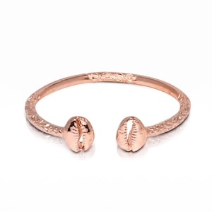 May include: A rose gold-toned open cuff bracelet featuring a polished finish. The band is decorated with an intricate engraved floral pattern along its length, and each end is capped with a metallic cowrie shell design facing inward.