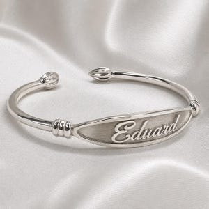 May include: A silver-toned open cuff bracelet featuring a central oval plaque with a brushed finish. The name Eduard is engraved in a cursive script on the plaque. The bracelet has ribbed detailing near the plaque and rounded, tapered ends.