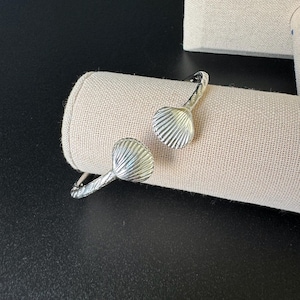 Könnte beinhalten: Silberarmband mit zwei Muschel-Anhängern. Das Armband ist offen und hat ein strukturiertes Design.