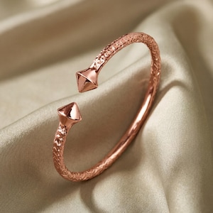 Peut inclure: Un bracelet manchette en or rose présentant une surface texturée et martelée le long de l'anneau, avec deux embouts lisses et polis en forme de pyramide aux extrémités ouvertes. Le métal a un éclat chaud et une structure rigide.