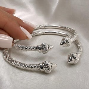 May include: Two silver-toned open cuff bracelets featuring a textured, embossed floral pattern along the band. Each end of the bracelets terminates in a polished, pointed, conical finial with vertical grooves, creating a distinct, reflective finish.