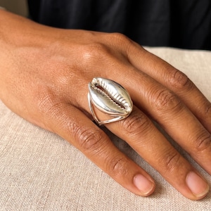 May include: A silver ring featuring a polished, elongated cowrie shell design. The metal surface is smooth and reflective, showcasing the natural, curved ridges and central opening characteristic of a cowrie shell, securely set on a simple band.