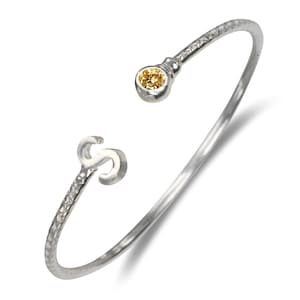 May include: A silver-toned open cuff bracelet with a textured band. The bracelet features a small, round, yellow gemstone at one end and a stylized letter "S" at the other. The bracelet is designed to be worn on the wrist.