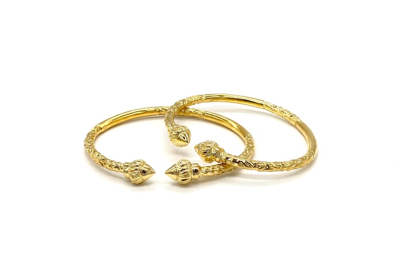 May include: Two gold-toned open bangles featuring a textured surface and pointed, cone-shaped finials at each end. The metal surface of the bracelets displays a repeating, intricate pattern along the entire length of the circular bands.