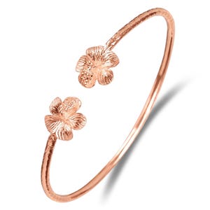 Könnte beinhalten: Ein Armreif aus Roségold mit einem strukturierten Band und zwei Hibiskusblütenmotiven an den offenen Enden. Das Metall hat eine polierte Oberfläche, die die detaillierten Blütenblätter und Staubgefäße der Blumen hervorhebt.