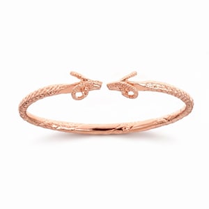 Könnte beinhalten: Ein offener Armreif aus Roségold mit zwei einander zugewandten Widderkopf-Endstücken. Das Armband hat eine texturierte, rindenartige Oberfläche, die sich von den Widderköpfen aus um den gesamten Umfang des runden Metallschmuckstücks erstreckt.