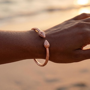 May include: A copper-colored cuff bracelet with a snake-head design. The bracelet features intricate detailing and is open at the wrist. The snake heads are at each end of the bracelet.