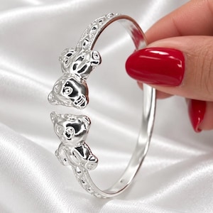 May include: A silver-toned open cuff bracelet featuring two sculpted bear heads at the ends. The band has a textured, diamond-patterned design along the outer surface, leading to the smooth, polished bear motifs that face each other.