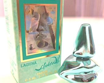 May include: A teal-colored perfume bottle and box set. The box features a gold-framed image of a face and the word "LAGUNA". The bottle is shaped like a pair of lips and a nose. The box reads "PARFUM e 5 ML - 0.17 US Fl Oz".