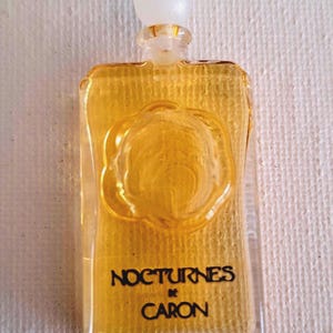 Puede incluir: Un frasco de perfume rectangular de vidrio lleno de un líquido dorado. El frasco presenta un emblema floral circular en relieve en la parte frontal y un tapón esférico blanco. El texto NOCTURNES DE CARON está impreso en negro en la parte inferior.