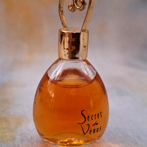 Può includere: Un piccolo flacone di profumo in vetro trasparente riempito di liquido ambrato. Il flacone ha un tappo color oro e un manico a forma di cuore. Le parole "Secret de Vénus" sono scritte in nero sul davanti.