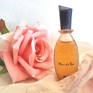 Może przedstawiać: Brzoskwiniowy flakon perfum z czarną nakrętką, z napisem "Fleur de Boa", stoi obok dużej różowej róży. Butelka ma dekoracyjny wzór róży i znajduje się na kremowej tkaninie z koronkowym wykończeniem.