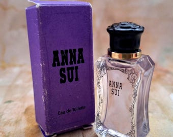 May include: A small, clear perfume bottle with a black cap and gold trim, next to a purple box. The bottle and box both feature the text "ANNA SUI". The box also has "Eau de Toilette" printed on it. The background is a light brown.