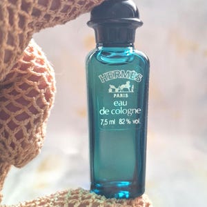 Puede incluir: Pequeña botella de perfume de color turquesa con tapón negro. La botella tiene una etiqueta con la marca "Hermès" y las palabras "Paris Eau de Cologne". Contiene 7,5 ml de líquido, 82% vol.