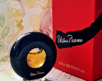 May include: A circular black perfume bottle with a central clear glass window revealing amber liquid, featuring the text Paloma Picasso in gold. Beside it stands a rectangular red box with a black brushstroke design and the text Paloma Picasso.
