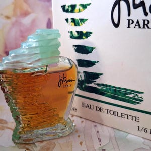 May include: A miniature glass perfume bottle with amber liquid and a tiered, light green cap. The bottle features the black text Jitrois Paris. Beside it stands a white box with green brushstroke accents, the text Jitrois Paris, and 1/6 FL.O.