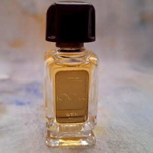 Può includere: Un piccolo flacone di profumo rettangolare con un tappo marrone scuro. La bottiglia è riempita con un liquido color oro e presenta la parola "WEIL" in un carattere color oro. La bottiglia ha una forma chiara e rettangolare.