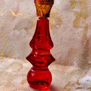 Könnte beinhalten: Ein kleiner, durchscheinender roter Glasparfümflakon mit abgestufter, bauchiger Silhouette und einem metallisch goldfarbenen Verschluss mit geprägten Details. Auf dem Boden des Flakons befindet sich der schwarze Text E2190CB.