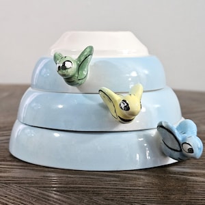 May include: A set of three blue ceramic bowls with bee-shaped handles. The bowls are stacked on top of each other, with the smallest bowl on top. The bee handles are painted in green, yellow, and blue.