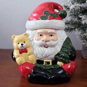May include: A ceramic Santa Claus cookie jar featuring a red hat with holly, a white beard, and a black belt. Santa holds a light brown teddy bear with a red bow on the right and a green Christmas tree on the left, wearing black boots.