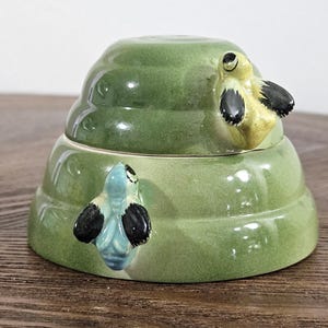 May include: A green ceramic beehive shaped sugar bowl with two bee figurines. The beehive has three tiers and the bees are perched on the top tier.