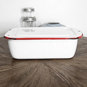 May include: A white rectangular enamelware container with a red trim around the rim. The container is sitting on a wooden surface. Two small glass jars with metal lids are in the background.