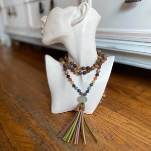Puede incluir: Un collar de cuentas con borla se exhibe sobre un busto blanco. El collar presenta una variedad de cuentas en tonos tierra, con una piedra verde y una borla con acentos rosas, verdes y marrones. El busto se encuentra sobre un fondo borroso.