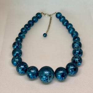 May include: A blue beaded necklace with a silver chain and clasp. The beads vary in size, with the largest ones in the center. The beads have a speckled, textured appearance. The necklace is displayed on a white surface.
