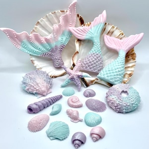May include: A set of pastel-coloured mermaid tail and seashell cake toppers. The tails are in shades of pink, purple, and blue, and the shells are in shades of pink, purple, and green. There is also a starfish cake topper.
