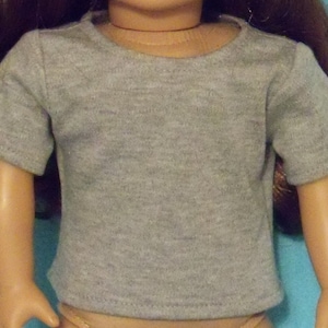 May include: A gray t-shirt with short sleeves, designed for a doll. The shirt has a crew neck and is made of a soft, heathered fabric. The doll's arms are visible, and the background is a solid blue.