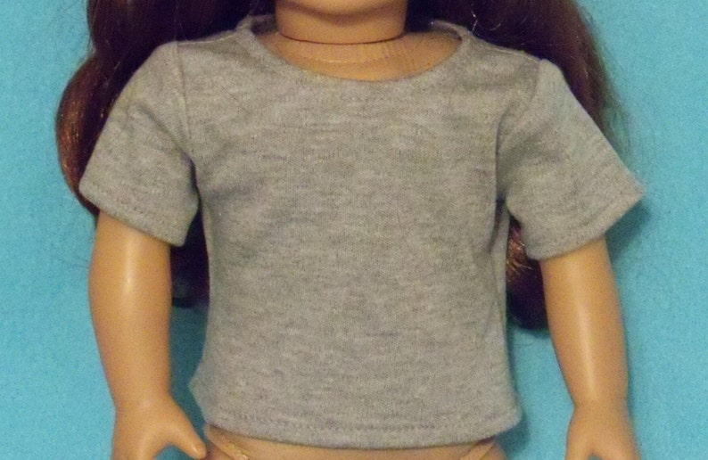 May include: A gray t-shirt with short sleeves, designed for a doll. The shirt has a crew neck and is made of a soft, heathered fabric. The doll's arms are visible, and the background is a solid blue.