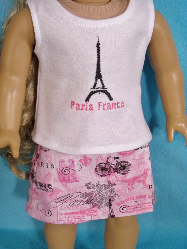 May include: A white sleeveless shirt with a black Eiffel Tower graphic and the text "Paris France" in pink. The shirt is paired with a pink skirt featuring a Parisian-themed print with bicycles, roses, and the word "Paris".