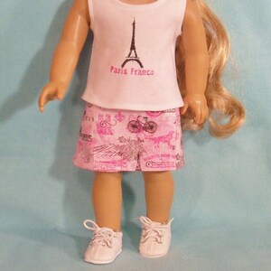 May include: A pink and white doll outfit with a white sleeveless shirt featuring a black Eiffel Tower graphic and the text "Paris France". The outfit also includes a pink skirt with a black and white Parisian-themed print.