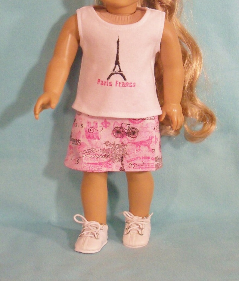 May include: A pink and white doll outfit with a white sleeveless shirt featuring a black Eiffel Tower graphic and the text "Paris France". The outfit also includes a pink skirt with a black and white Parisian-themed print.