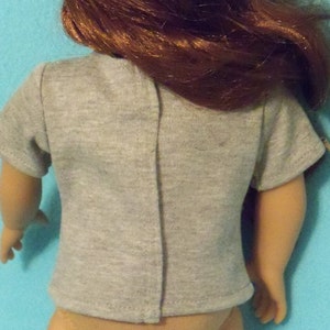 May include: A gray t-shirt with short sleeves, designed for a doll. The back of the shirt is visible, showing a vertical seam down the center. The doll has auburn hair and is against a light blue background.
