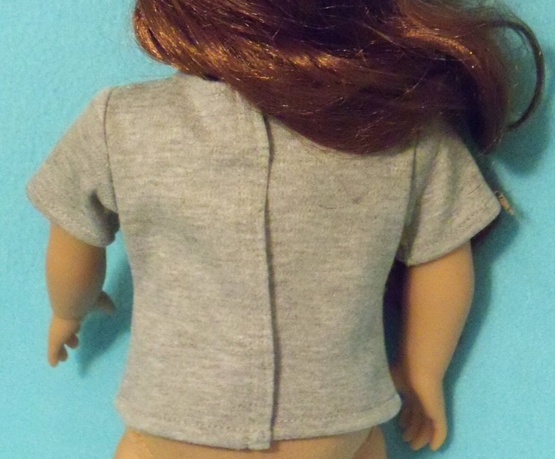 May include: A gray t-shirt with short sleeves, designed for a doll. The back of the shirt is visible, showing a vertical seam down the center. The doll has auburn hair and is against a light blue background.
