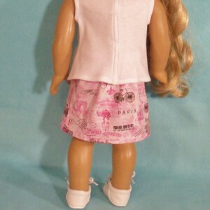 May include: A pink and white doll outfit with a white sleeveless top and a pink skirt with a Parisian print. The skirt has the word "Paris" printed on it multiple times.
